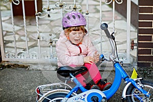 Cute little girl sitting on the ground after falling off her bike. Upset crying preschool child with safe helmet getting