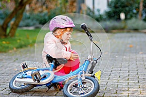 Cute little girl sitting on the ground after falling off her bike. Upset crying preschool child with safe helmet getting