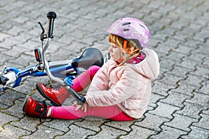 Cute little girl sitting on the ground after falling off her bike. Upset crying preschool child with safe helmet getting