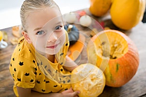 Cute little girl sitting on kitchen table, helping to carve large pumpkin, looking at camera and smiling. Halloween. photo