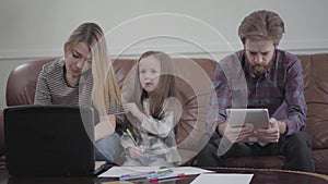 Cute little girl sitting between mother and father drawing with colored markers. Young woman typing on her laptop