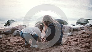 Cute little girl sitting on shore and playing takes sand in hand and then pours on the ground. Brother going to sister.