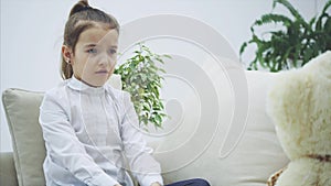 Cute little girl sitting on the sofa, talking with her teddy. It doesn't understand her. She looks disappointed.