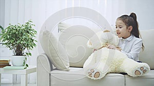 Cute little girl sitting on the sofa, talking with her teddy, whispering some secret in its ear.