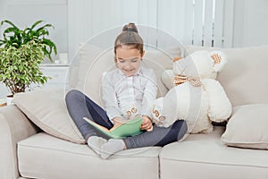 Cute little girl sitting on the sofa with teddy-bear near her, reading fairy-tale, pointing her finger at something in
