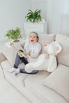 Cute little girl sitting on the sofa with teddy-bear near her, reading fairy-tale, pointing her finger at something in