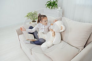 Cute little girl sitting on the sofa with teddy-bear near her, reading fairy-tale, using different gestures to explain