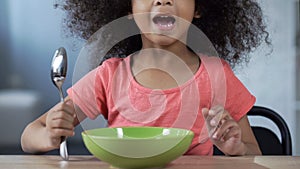 Cute little girl sitting at table with spoon and asking for dinner, hungry kid