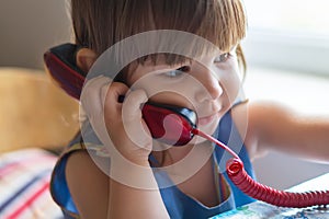 A cute little girl is sitting at a table and talking on an old red home landline phone. Communication, the concept of relationship