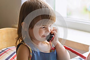 Cute little girl sitting at the table and talking on an old red home phone. Communication, the concept of relationships. European