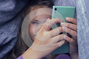 Cute little girl sitting under blanket with phone.