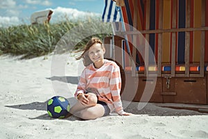 Cute little girl sitting on white sand with ball on beautiful beach with dunes on Baltic sea