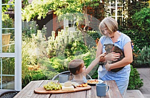 Cute little girl sittting at table and having breakfast in garden of country house and petting cat