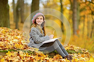 Cute little girl sketching outside on beautiful autumn day. Happy child playing in autumn park. Kid drawing with colourful pencils
