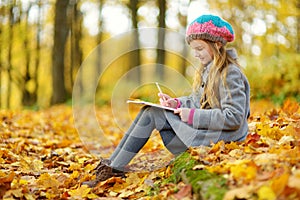 Cute little girl sketching outside on beautiful autumn day. Happy child playing in autumn park. Kid drawing with colourful pencils