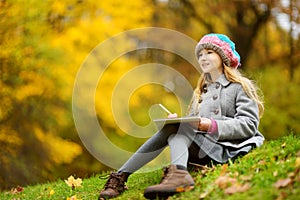 Cute little girl sketching outside on beautiful autumn day. Happy child playing in autumn park. Kid drawing with colourful pencils