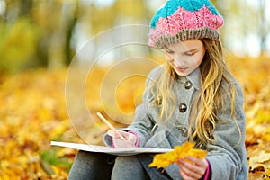 Cute little girl sketching outside on beautiful autumn day. Happy child playing in autumn park. Kid drawing with colourful pencils