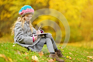 Cute little girl sketching outside on beautiful autumn day. Happy child playing in autumn park. Kid drawing with colourful pencils