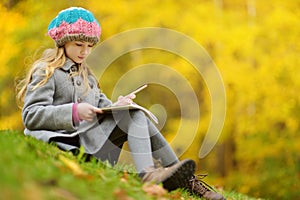 Cute little girl sketching outside on beautiful autumn day. Happy child playing in autumn park. Kid drawing with colourful pencils