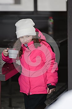 Cute little girl in ski suit drinking hot tea from cup during cold winter outdoor leisure activity on mountain resort