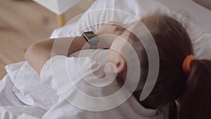 Cute little girl sleeping in the bed at home. Adorable girl waking up from an alarm clock on her smart watch. Modern