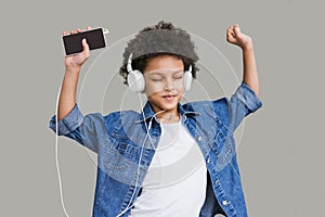 Cute little girl with smartphone and headphones listening to music and dancing. Isolated on grey background studio shot