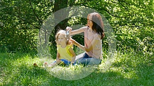 Cute Little Girl is Smiling while Beautiful Young Mother is Combing Her Hair Sitting on the Green Grass in the Park. Mom