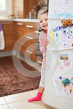 Cute little girl smiling behind refrigerator door in kitchen