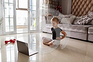 Cute little girl in sportswear watching online video on laptop and doing fitness exercises at home. Distant training with personal