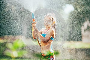 Cute little girl sprinkls a water for herself from the hose, makes a rain. pleasure for hot summer days