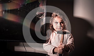 Cute little girl standing in front of a TV with broken screen holding a hammer. Home insurance concept