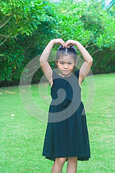 Cute little girl standing on green grass and raise up hers hand to make heart shape over head with sunlight background.