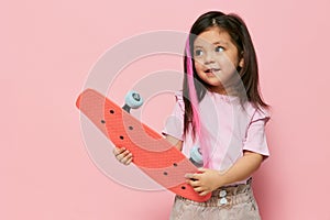 a cute little girl is standing on a pink background in a pink T-shirt, holding her skate in her hands, looking at it