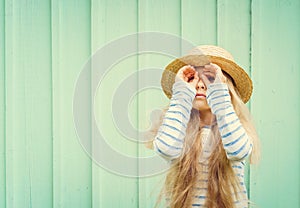 Cute little girl stands near a turquoise wall in boater hat and looks invented binoculars. Space for text