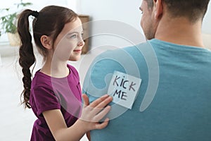 Cute little girl sticking note with words Kick Me to father`s back at home photo