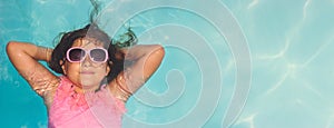 Cute little girl with sunglasses, smiling and laying relaxed in the pool; summertime background with copy space