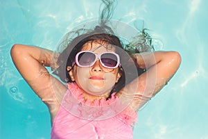 Cute little girl with sunglasses, smiling and laying relaxed in the pool; summertime background with copy space