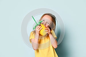 Cute little girl in sunny yellow t-shirt making a show of biting bright inflatable pineapple. Waist up shot isolated on light blue