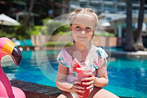 Cute little girl in swimsuit sitting near swimming pool with inflatable pink flamingo and drinking fresh watermelon juice. Summer