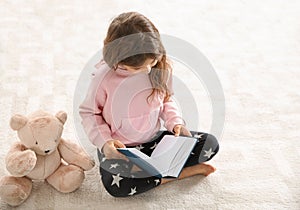 Cute little girl with teddy bear reading book on floor.