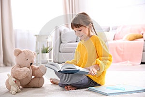 Cute little girl with teddy bear reading book on floor