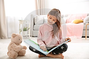 Cute little girl with teddy bear reading book on floor