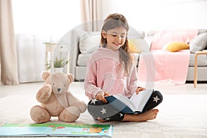 Cute little girl with teddy bear reading book on floor