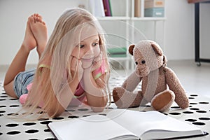 Cute little girl with teddy bear reading book on floor at home