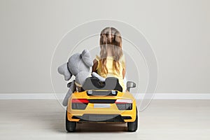 Cute little girl with toy elephant driving children`s car near grey wall indoors, back view