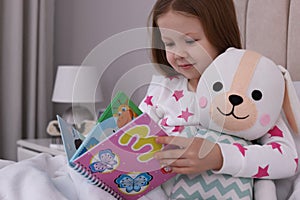 Cute little girl with toy reading book in bed at home