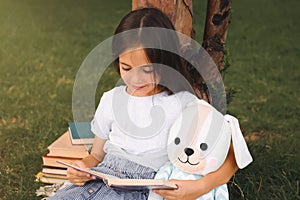 Cute little girl with toy reading book on green grass near tree