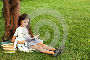 Cute little girl with toy reading book on green grass near tree in park