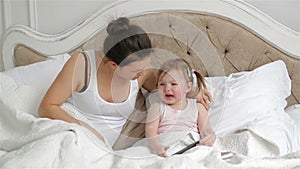 Cute Little Girl with Two Ponytails is Crying Sitting on the Bed. Mother is Trying to Calm Her Daughter in the Bedroom.