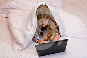 Cute little girl using a laptop in bed under a blanket